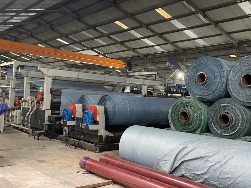 Mesin laminasi terpal dengan tumpukan roll hijau dan biru di pabrik Yo Tarpaulin